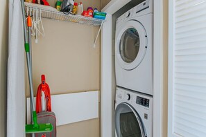 Ein Waschraum mit einer gestapelten Waschmaschine und einem Trockner, einem Mop, einem Besen und einem Regal mit Reinigungsmitteln.