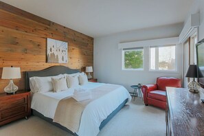 Un dormitorio con cabecera de madera, una cama grande con sábanas blancas, un sillón de cuero rojo, una mesita y una ventana con vista a follaje.