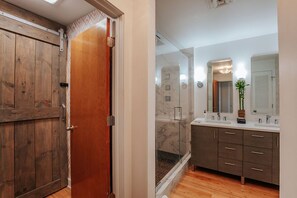 Un baño moderno con puerta de madera, cabina de ducha de vidrio y tocador con varios lavamanos y cajones.