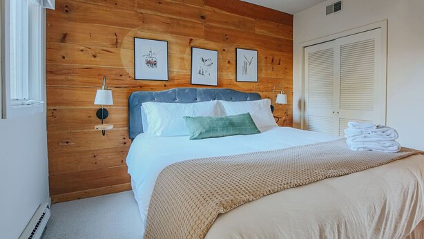 Een slaapkamer met een wand van houten panelen, een bed met wit beddengoed, twee wandlampen en een deur met verstelbare luiken.