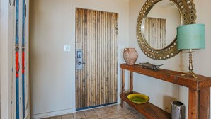 Een kamer met een houten deur, een spiegel met decoratieve lijst, een houten console en een lamp, plus een klein plankje met decoratieve objecten.