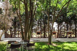 An outdoor seating area with tables and chairs, surrounded by trees and a barbecue grill.