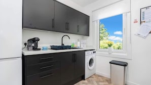A compact kitchen with dark cabinets, a washing machine, a sink, and a window with a view of greenery. - Fraser Apartment- 2 Bedrooms Inverleith, Edinburgh (Edinburgh)
