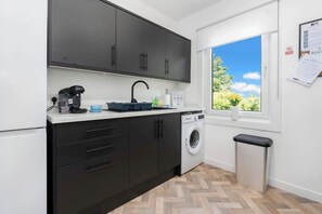 Eine kleine Küche mit dunklen Schränken, einer Waschmaschine, einem Spülbecken und einem Fenster mit Blick auf Grünflächen.