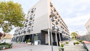 Un immeuble moderne à plusieurs étages, doté de balcons, d’une entrée couverte et d’un trottoir pavé agrémenté de plantes en pot.