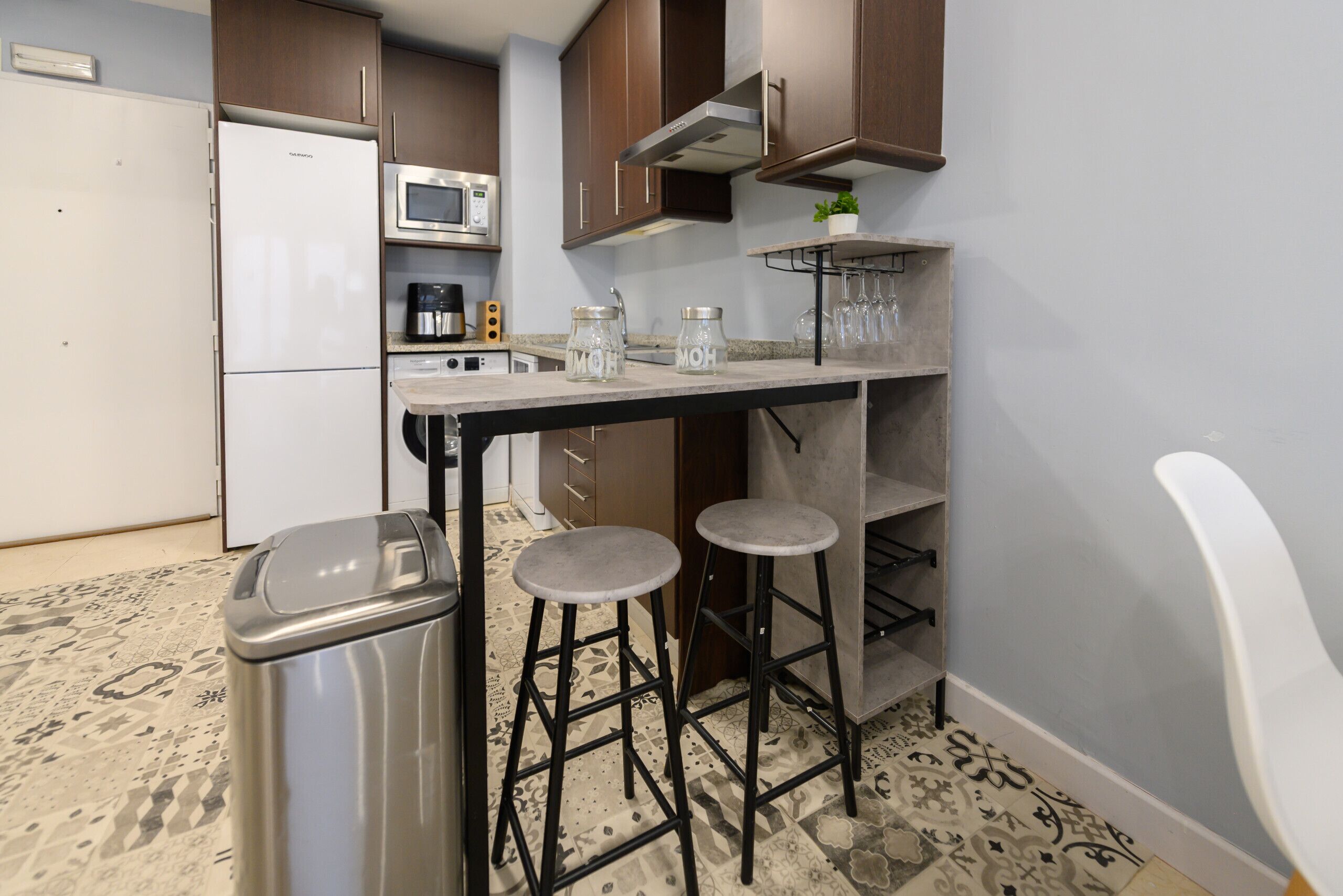 Una cocina compacta equipada con refrigerador, microondas y cafetera. Hay dos taburetes y un cubo de basura.