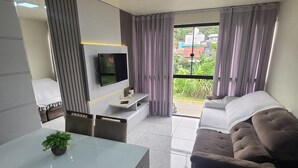 A modern living room with a flat-screen TV mounted on the wall, a sofa, a dining table with chairs, and a balcony with curtains. - ER21C - Bombinhas Center Residential (Bombinhas)