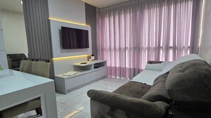 A modern living room with a flat-screen TV mounted on the wall, a sofa, a coffee table, and a dining area with a table and chairs. - ER21C - Bombinhas Center Residential (Bombinhas)