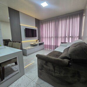 A modern living room with a flat-screen TV mounted on the wall, a sofa, a coffee table, and a dining area with a table and chairs. - ER21C - Bombinhas Center Residential (Bombinhas)