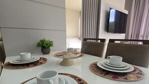 A dining area with a white table set for four, including plates, cups, and a small decorative bowl. There is a potted plant on the table, and a television mounted on the wall in the background. - ER21C - Bombinhas Center Residential (Bombinhas)
