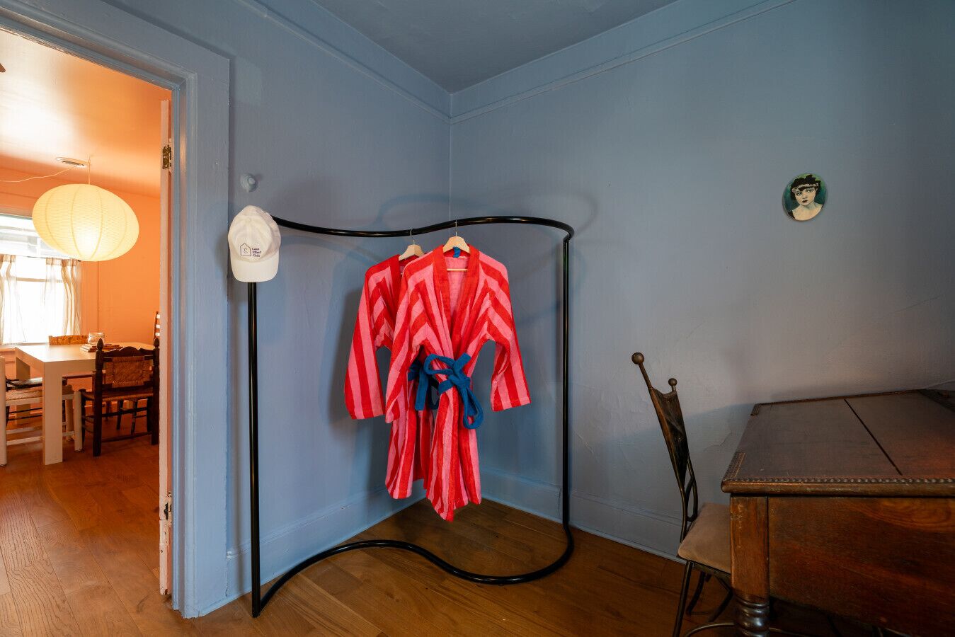 Una habitación con una pared azul, suelo de madera, zona de comedor con mesa y sillas, y un perchero con una bata de baño a rayas rojas y blancas.