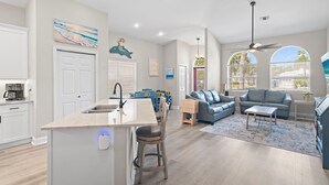Una cocina moderna con una isla central, un comedor con sillas azules, una sala de estar con sofá y mesa de centro, y un ventilador de techo.