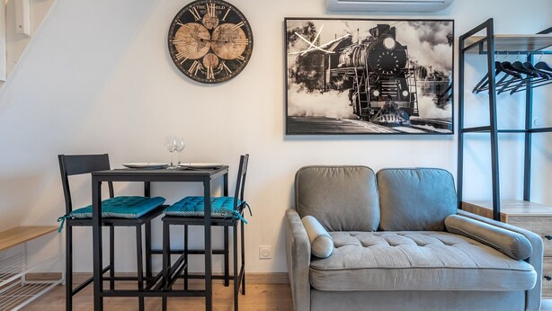 A living room with a grey sofa, a dining table with chairs, a wall clock, and a framed picture of a train. - Au Bonheur Du Voyageur - Proche gare routière (Vannes)