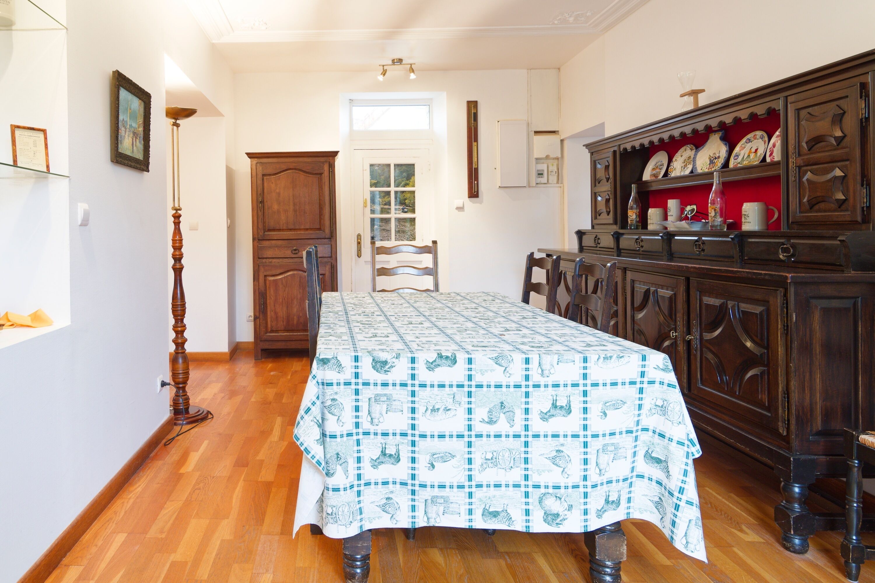 Uma área de refeições com uma mesa coberta por uma toalha estampada, cadeiras de madeira, um armário com pratos decorativos e piso de madeira.