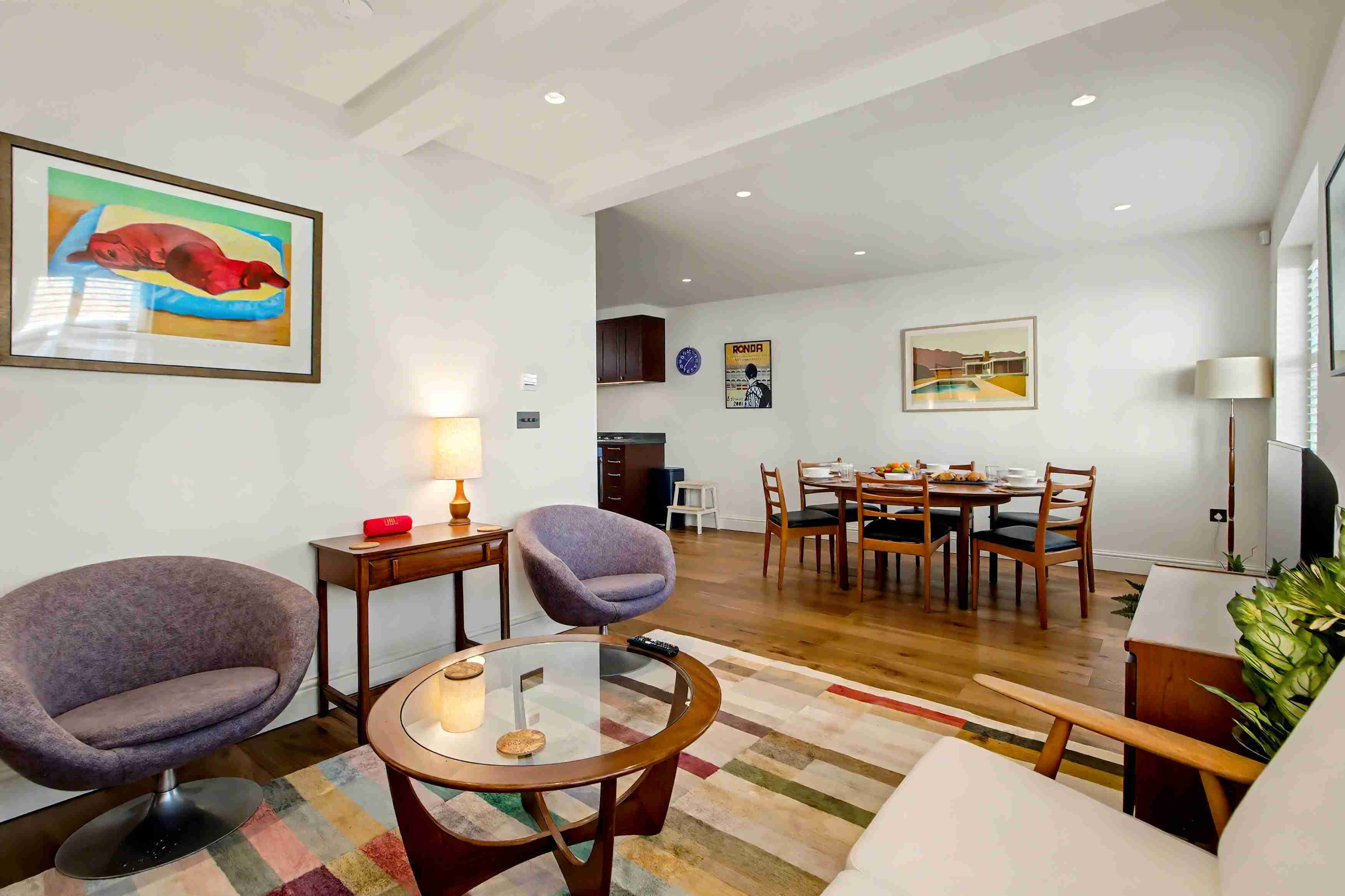 A modern living room with a glass coffee table, two armchairs, a dining area with a wooden table and chairs, and a kitchen in the background.