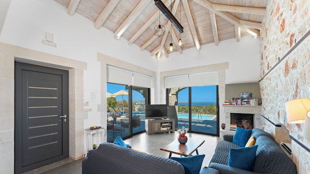 A spacious living room with a large sofa, a coffee table, and a television. There is a fireplace and a bookshelf. The room has large windows with a view of the ocean. - Villa Resi, Vamos, Private Pool, Sea View-5 minutes to the beach! (Chania)