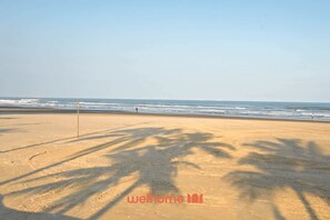 Ein Strand mit dem Schatten einer Palme, einem einzelnen Pfahl und dem Meer im Hintergrund.