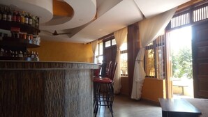 A bar area with a wooden counter, red bar stools, and a shelf with bottles. There are large windows with curtains and a view of greenery outside. - Northern Galaxy Hotel Isiolo (Isiolo)