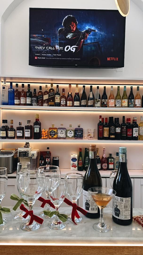 A bar area with a television displaying a movie poster, shelves of liquor bottles, and glasses on the counter.
