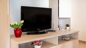 Una sala de estar moderna con un televisor de pantalla plana, un jarrón rojo con flores blancas, una planta pequeña en maceta y un objeto decorativo de cristal sobre una consola de madera.