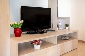 Ein modernes Wohnzimmer mit einem Flachbildfernseher, einer roten Vase mit weißen Blumen, einem kleinen Topfpflanzen und einem dekorativen Glasobjekt auf einem Holztischelement.