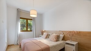 A bedroom with a bed, bedside tables, a nightstand, a window with curtains, and a hanging light fixture.