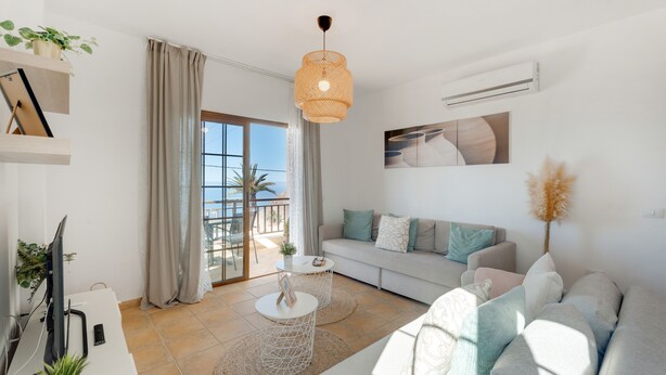 A modern living room with a sofa, a coffee table, a hanging light fixture, and a view through a glass door to an outdoor balcony.