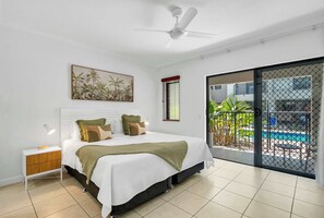 Ein Schlafzimmer mit Bett, Nachttisch, Deckenventilator, Kunstwerk, Fenster und Blick auf einen Außenpool.