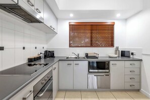 Eine moderne Küche mit Edelstahlherd, Backofen, Mikrowelle und Geschirrspüler. Ein Fenster mit Holzjalousien, eine Spüle und eine Arbeitsplatte mit Wasserkocher und Geschirrgestell.