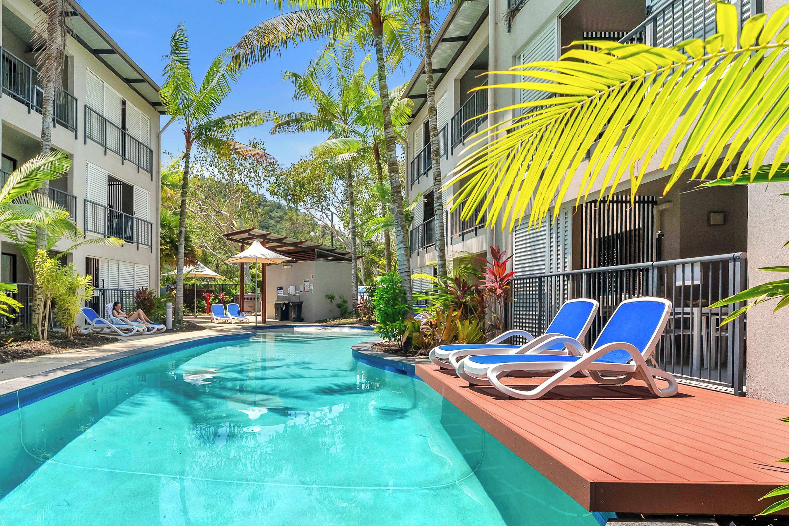 A swimming pool with lounge chairs, surrounded by buildings and palm trees.