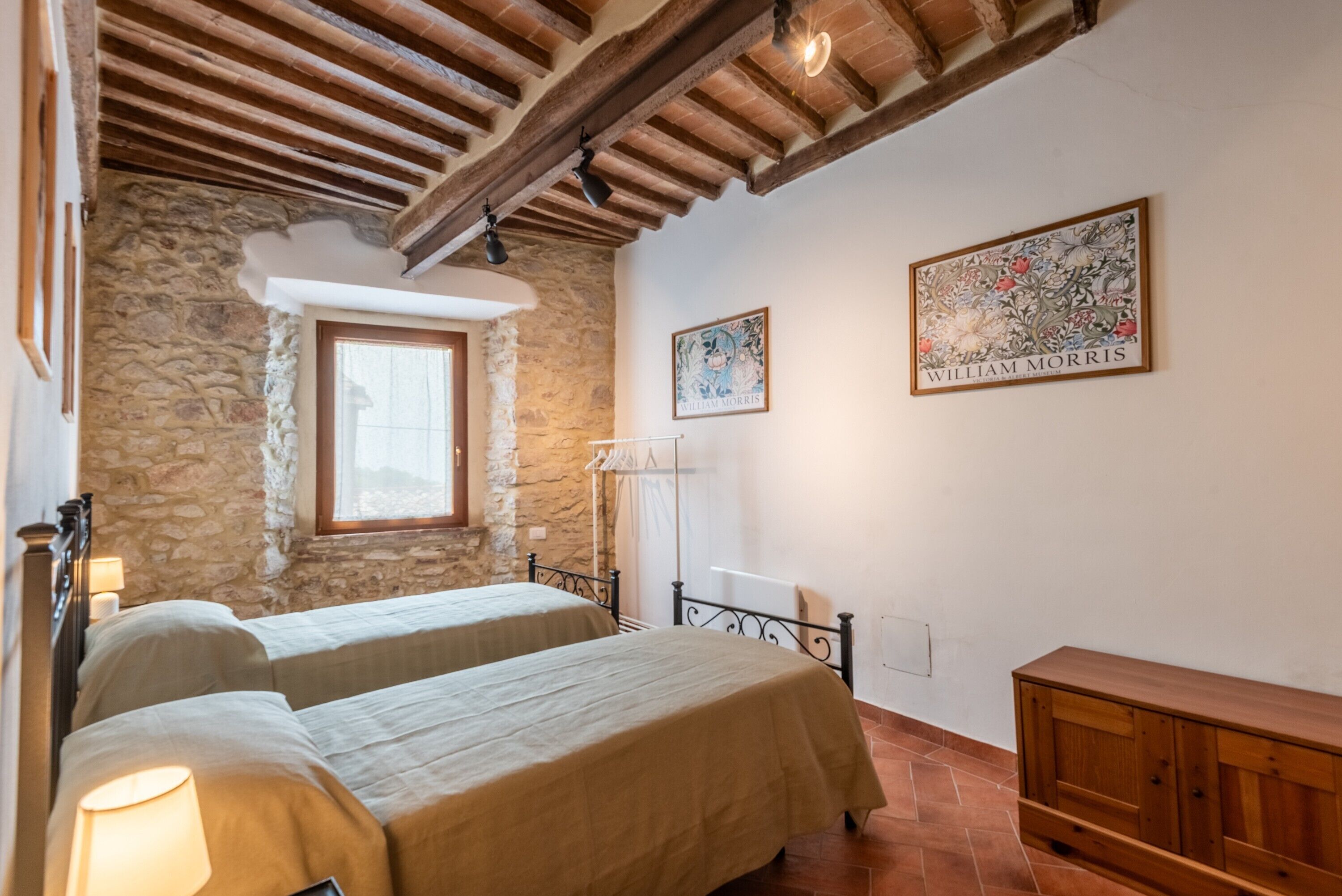 Une chambre avec deux lits, des murs en pierre, des poutres en bois, une fenêtre et des œuvres d’art.
