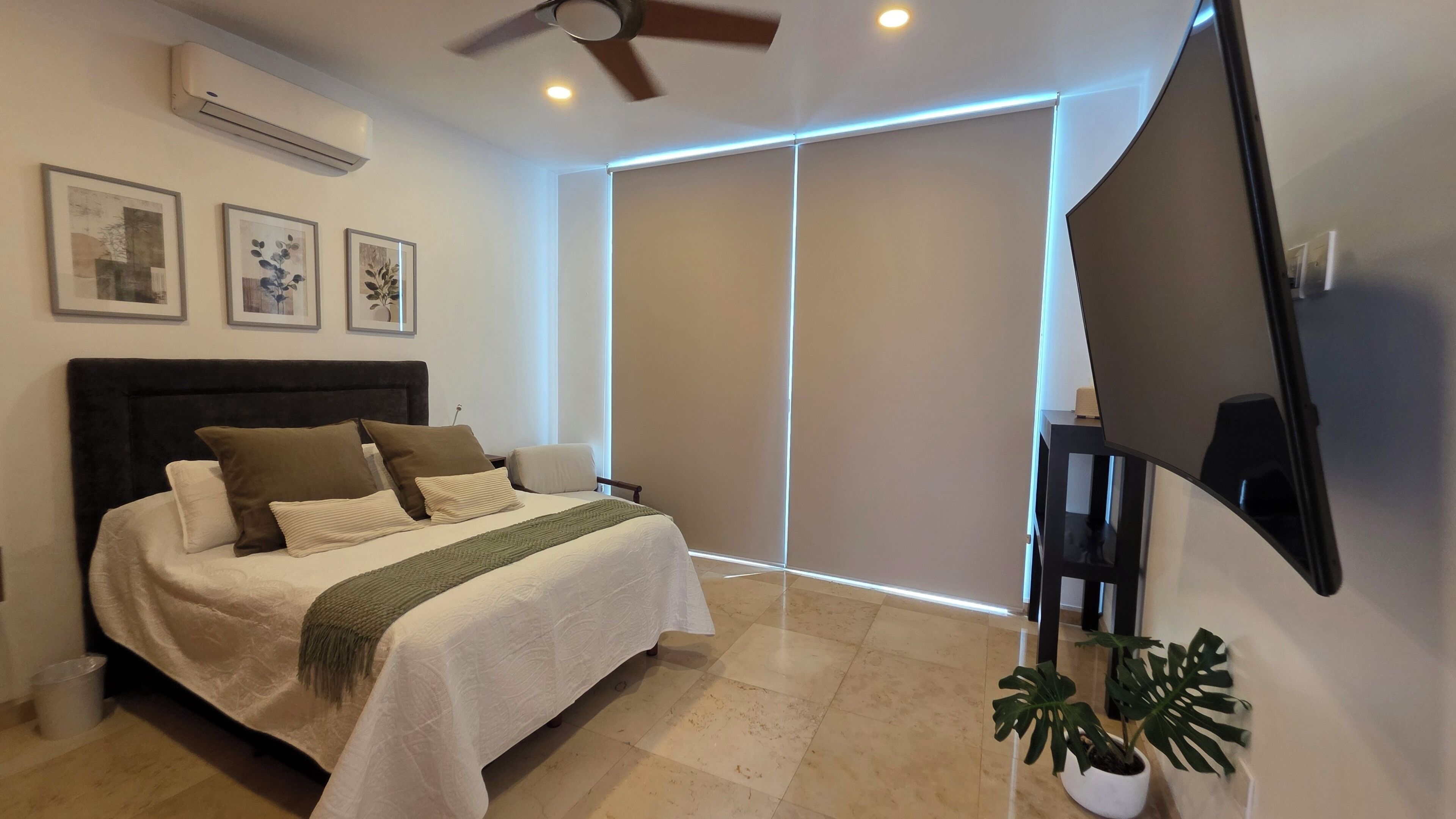 A bedroom with a bed, pillows, a blanket, a ceiling fan, a TV, and a plant.