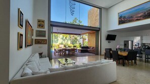 Una sala de estar moderna con un amplio ventanal que da a un patio exterior, un comedor y una cocina a la vista a través del vidrio.