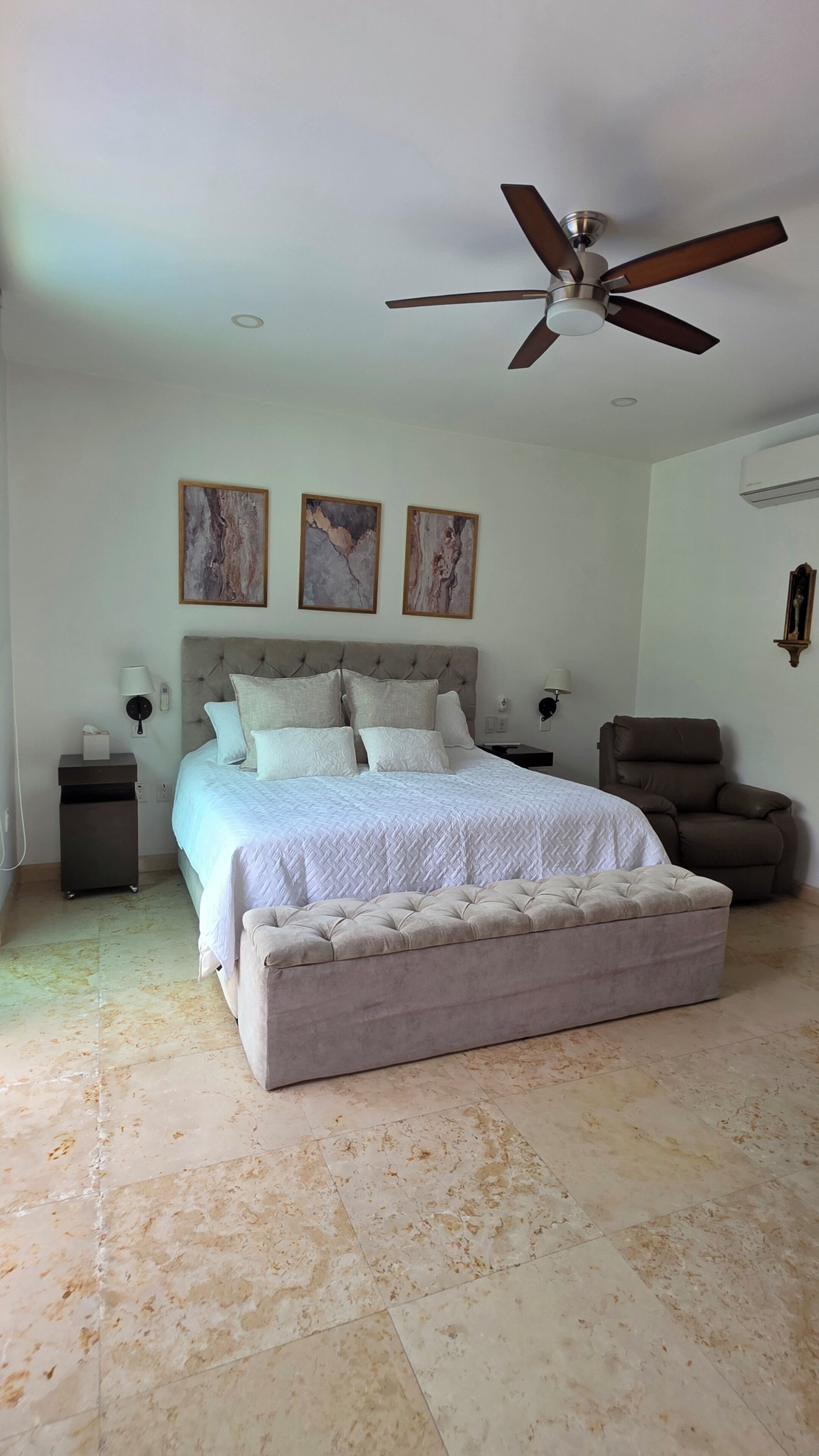 Un dormitorio con cama, ventilador de techo, una silla y obras de arte en la pared.