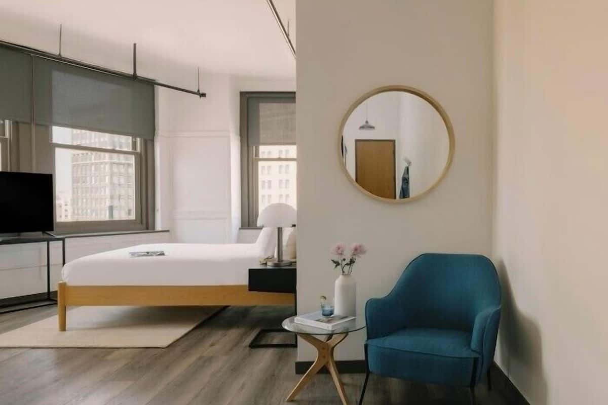 A modern hotel room with a bed, a round mirror, a blue armchair, and a small table with a vase.