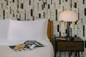Una cama bien hecha con una revista y un bolígrafo encima, una lámpara sobre una mesita de noche y una pared con estampado de periódico.