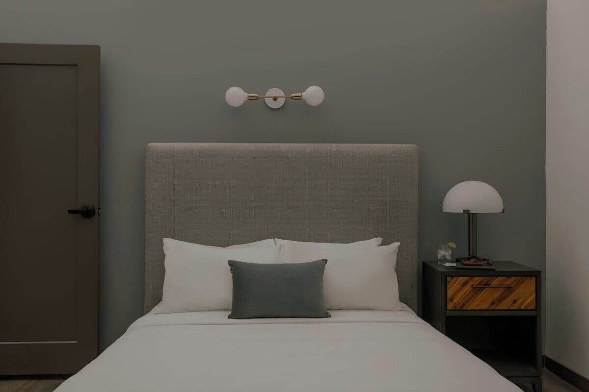 A neatly made bed with white and gray bedding, a gray headboard, and a bedside table with a lamp and a small decorative item.