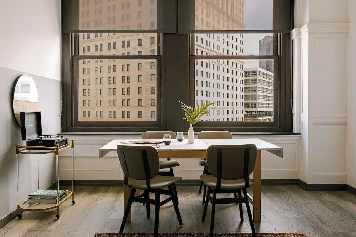 A modern dining area with a large window, a dining table, four chairs, a small plant, and a bookshelf.