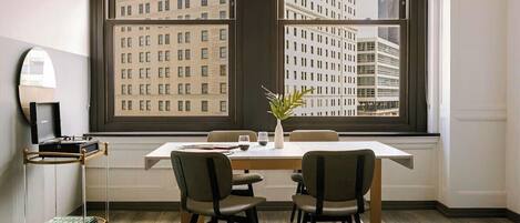 A modern dining area with a large window, a dining table, four chairs, a small plant, and a bookshelf.