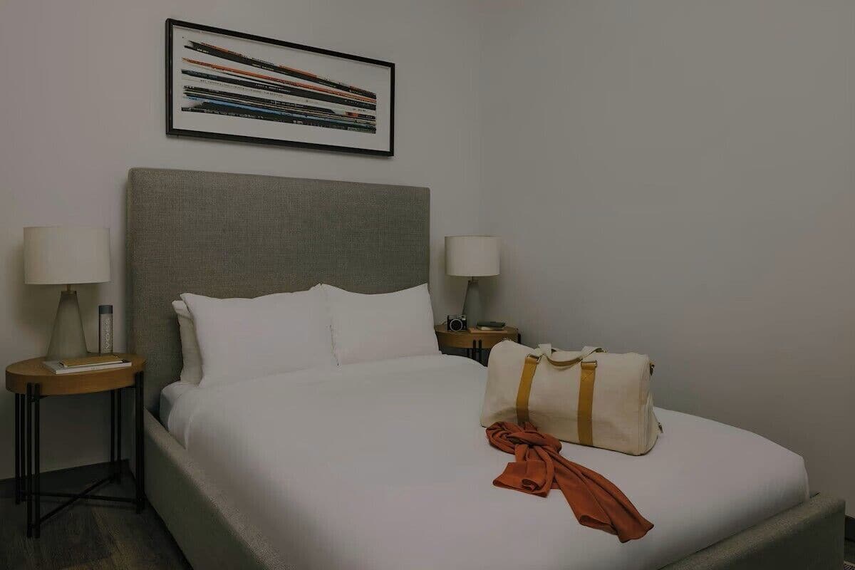 A neatly made bed with white linens, a grey headboard, and a framed picture above it. There are two bedside tables with lamps and a bag on the bed.