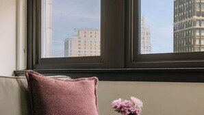 Een moderne woonkamer met een grijze bank, een roze kussen, een klein houten bijzettafeltje met een bloemenvaas, en een koffiemok op een glazen tafel.
