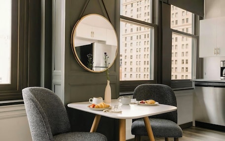 A modern dining area with a round mirror, two grey upholstered chairs, a white table, and a view of tall buildings through large windows.