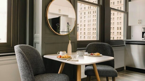 Een moderne eetruimte met een ronde spiegel, twee stoffeerde grijze stoelen, een witte tafel en een uitzicht op hoge gebouwen door grote ramen.