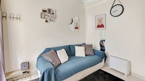 A modern bedroom with a blue sofa, white and grey cushions, a wall clock, and a radiator. - Charming Marais Historic Studio (Paris)