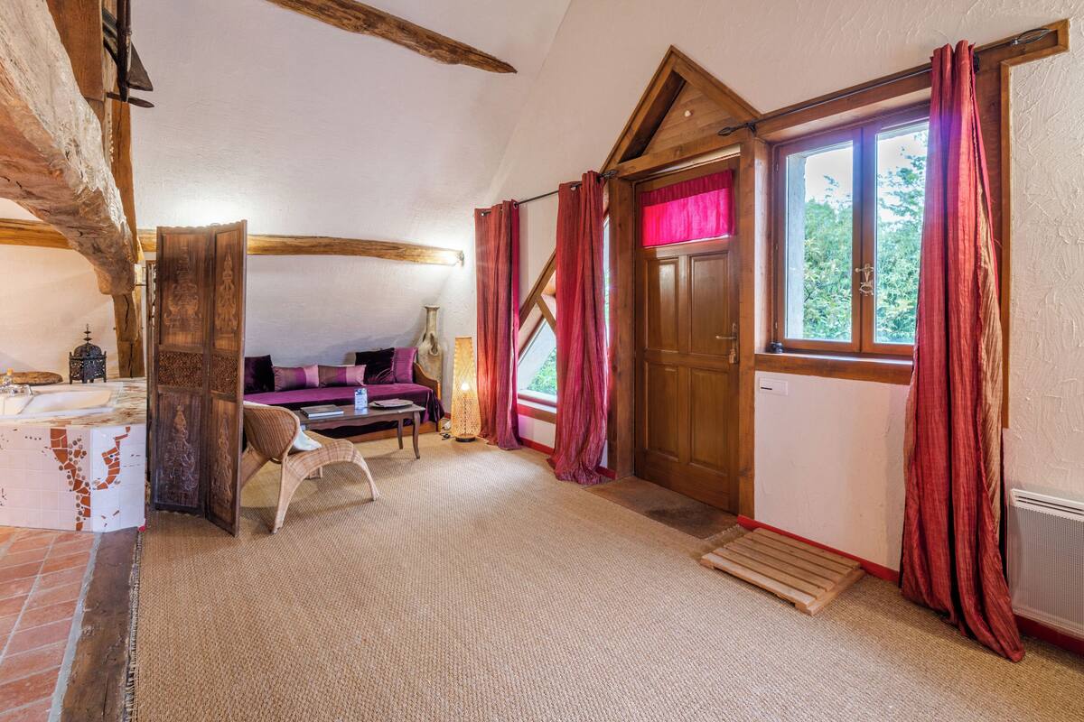 Una habitación acogedora con techo de madera, un ventanal con cortinas rojas, una puerta de madera y una pequeña mesa con una silla.