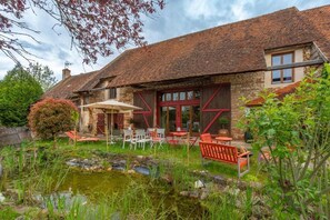 Un bâtiment rustique en pierre, doté d’un toit en tuiles, d’un espace extérieur aménagé avec des tables et des chaises, et d’un petit étang.