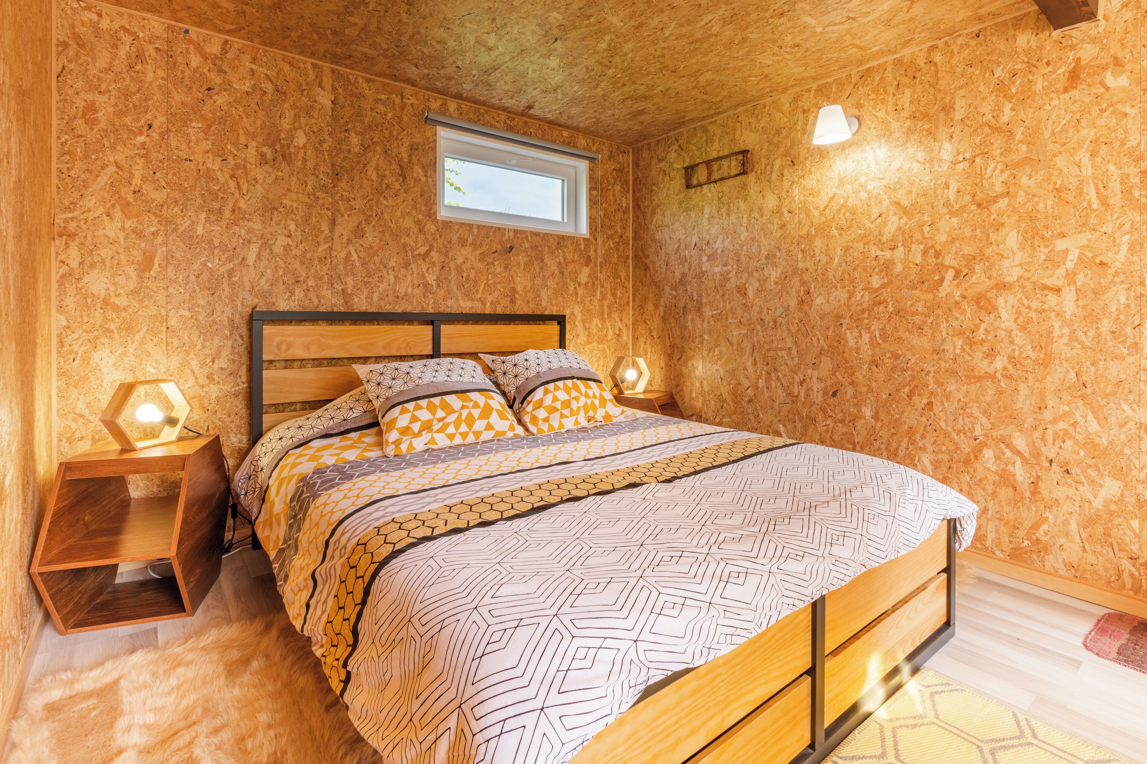 A wooden-paneled bedroom with a bed, a window, and a nightstand with a lamp.