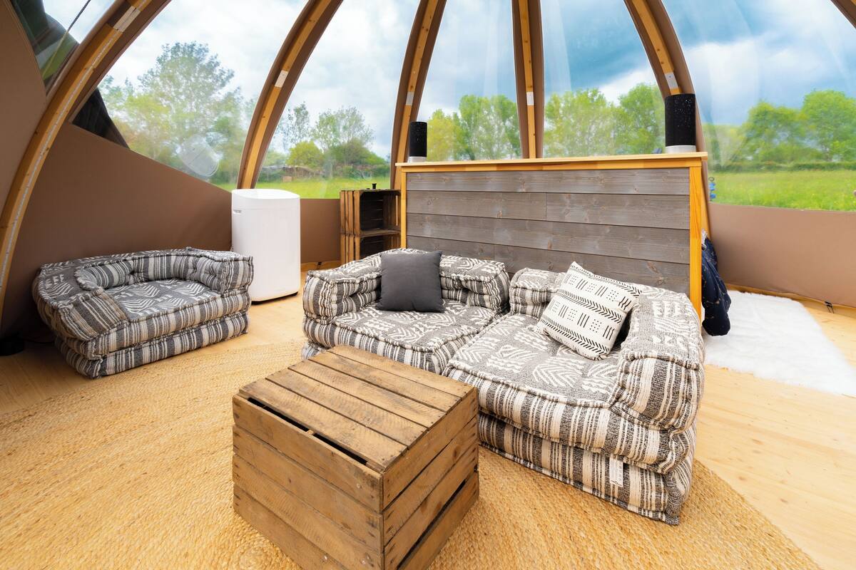 Un interior acogedor con un conjunto de sofá, una mesa de centro de madera y un electrodoméstico blanco, todo frente a un amplio ventanal arqueado con vistas a un paisaje verde.