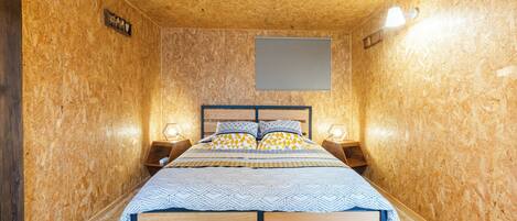A bedroom with a wooden headboard, a bed with patterned bedding, two bedside tables with lamps, and a wall-mounted light fixture.
