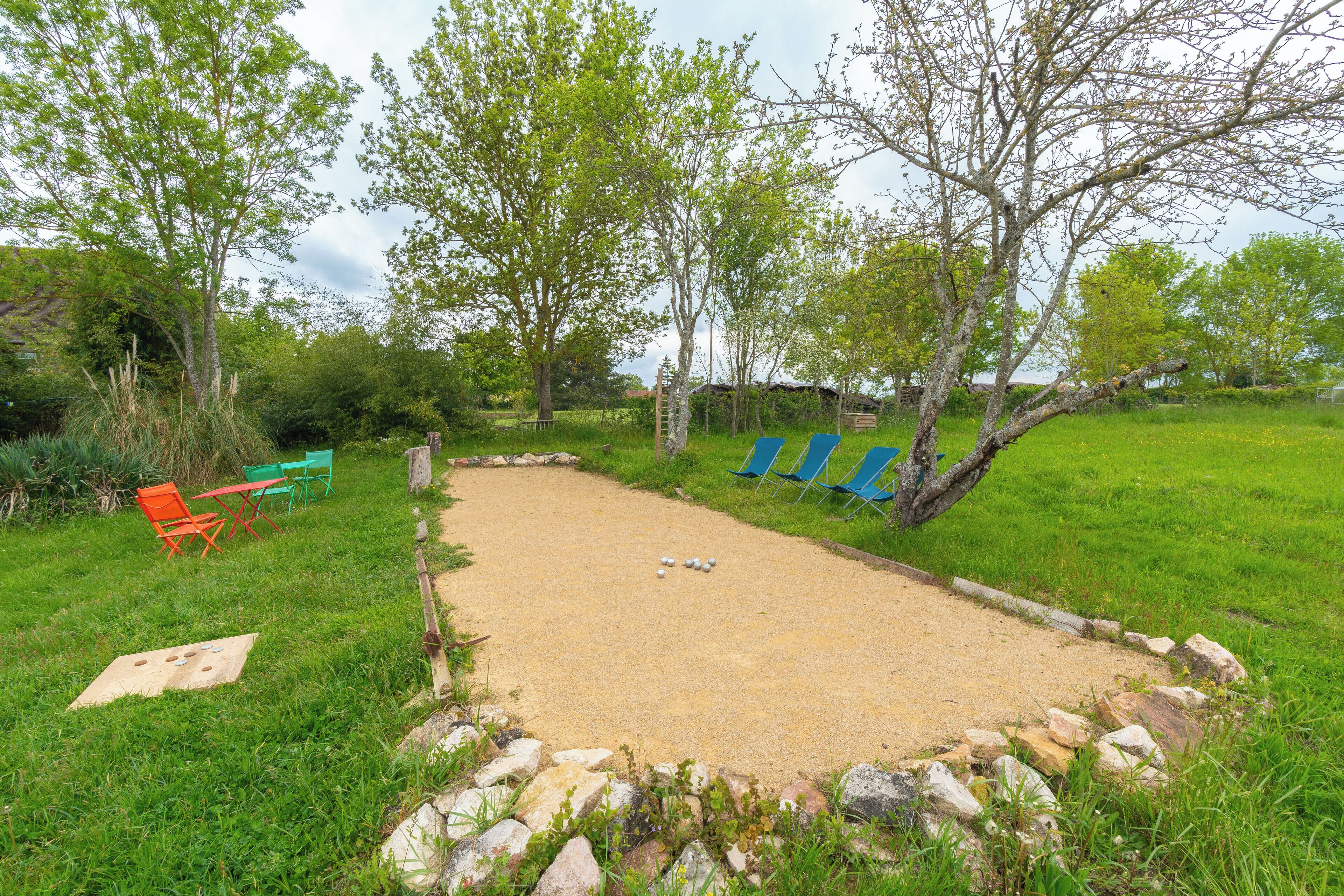 Una pista de terra baixa, una zona enjardinada, tot envoltat d'arbres i mobiliari per a l'exterior.